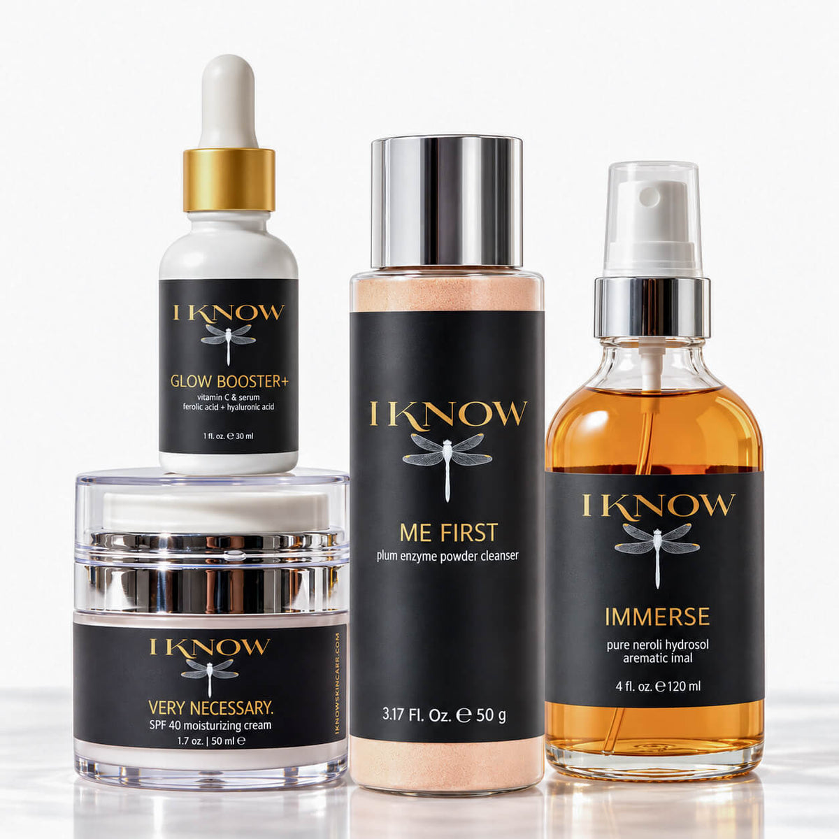 Four skincare products from IKnow Cosmetics arranged on a white background: a white dropper bottle with a gold cap labeled Glow Booster+, a beige powder bottle with a silver cap labeled Me First, a clear jar with a white lid labeled Very Necessary, and an amber spray bottle labeled Immerse.